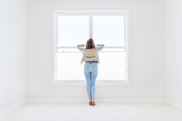 Fototapeta na wymiar Back of one young woman standing in white, bright empty, clean room on carpet of modern new house, home, apartment looking through the sunny large window with blinds