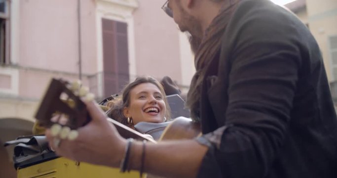 Man smiling while playing guitar music with girlfriend.Woman listening to boyfriend singing song.Caucasian couple in love roadtrip vacation italian travel on convertible vintage car.4k handheld video