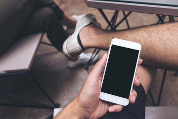 Mockup image of hand holding white mobile phone with blank black screen on thigh with white canvas shoes in cafe