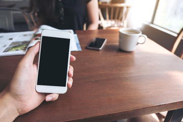 Mockup image of a man's hand holding white mobile phone with blank black screen , white coffee cups and woman reading book in modern loft cafe