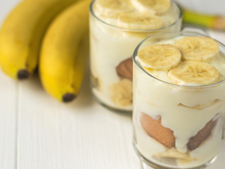 Two glass cups with banana pudding and two ripe bananas on a white table.