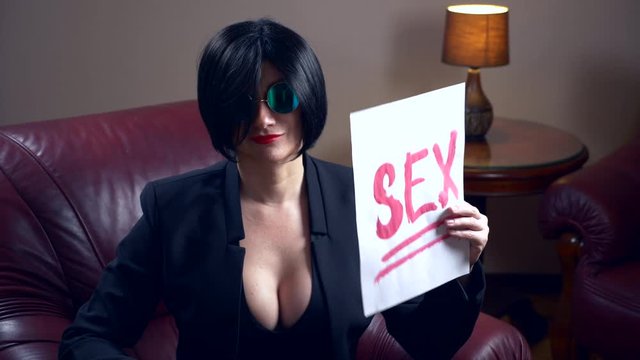 Hot brunette in round sunglasses, sits in an armchair and shows a sheet with the inscription sex. 4k, slow motionts