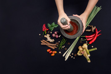 The Art of Thai Cuisine - Thai lady’s hands hold stone granite pestle with mortar and red curry paste ingredient together with fresh herbs and spices on classic dark background at top view angle.