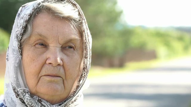 Thoughtful look of serious mature elderly woman aged 80s with gray hair dressed in a handkerchief on the background of road in summer. Close-up