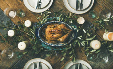 Flat-lay of holiday table setting for party, friends gathering or family celebration with oven roast chicken, plates, silverware, candles and olive branch decoration over rustic wooden table