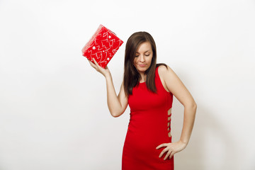 Interested and engaged young woman dressed in red dress and Christmas hat holds gift box, thinks that she was given on white background. Santa girl with present isolated. New Year holiday 2018 concept