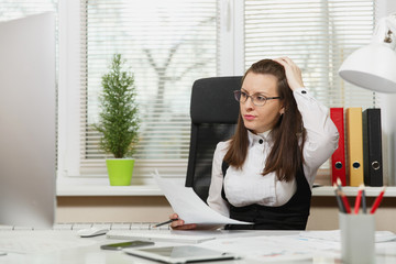 Beautiful serious and engrossed brown-hair business woman in suit and glasses sitting at the desk...