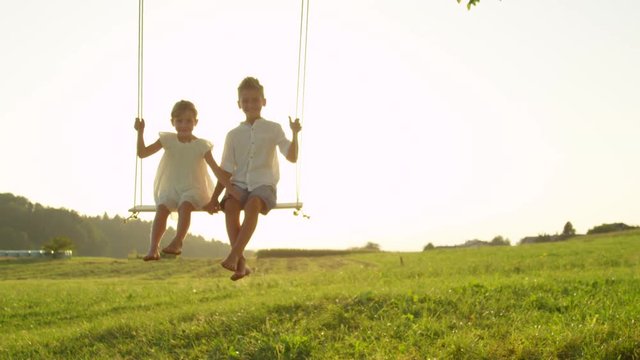 SLOW MOTION SILHOUETTE: Happy young brother and sister enjoying sunset on a wooden swing. Smiling little boy and girl swaying in the warm golden sunlight. Barefoot kids playing on a swing set at dusk