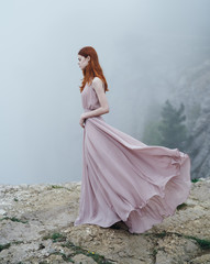 Fototapeta na wymiar Young beautiful woman in a long pink dress is standing on a cliff of high rock in the fog