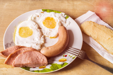 Fried eggs lie in a plate. Sandwich and sausage on a wooden background.