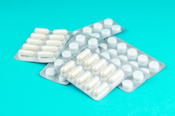 Packs of white capsules and pills packed in blisters with copy space on turquoise background. Focus on foreground, soft bokeh. Pharmacy drugstore concept