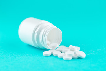 Close up white pill bottle with spilled out pills on turquoise background with copy space. Focus on foreground, soft bokeh. Pharmacy drugstore concept
