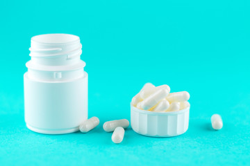 Close up white pill bottle with spilled out pills and capsules in cap on turquoise background with copy space. Focus on foreground, soft bokeh. Pharmacy drugstore concept