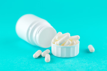 Close up white pill bottle with spilled out pills and capsules in cap on turquoise background with copy space. Focus on foreground, soft bokeh. Pharmacy drugstore concept