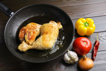 Two roasted chicken legs in black frying pan with vegetables and spices on dark wooden planks