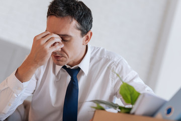 Painful thoughts. Emotional sad young man sitting at home with his eyes closed and touching his forehead while thinking about losing his favorite job