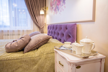 white tea set on the bedside table in  cozy bedroom with purple and gold textiles