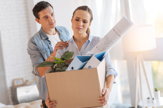 No Worries. Emotional Tired Sad Woman Carrying A Big Box Of Personal Items After Being Fired From Work While Her Kind Attentive Husband Persuading Her Not To Worry