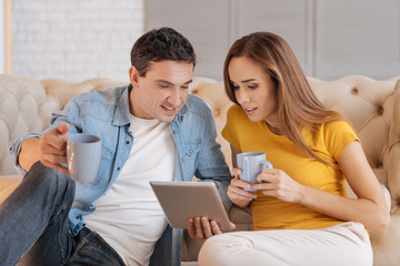 Exciting film. Cheerful loving couple sitting at home and drinking hot tasty tea while holding a tablet and watching an exciting film