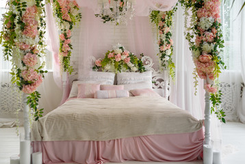 Honeymoon suite with canopy bed, free space. Luxurious wood canopy bed with flowers and pillows on it. Female bedroom in pink and white colors, copy space. Big comfortable bed in elegant bedroom