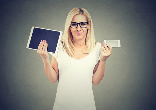 Perplexed woman in glasses shrugging shoulders uncertain what to choose tablet or smartphone 