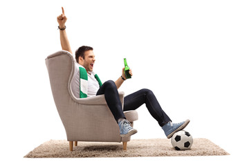Fototapeta Excited football fan with a scarf and a beer bottle seated in an armchair pointing up obraz