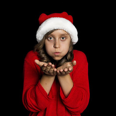 A young girl in a red sweater and a Santa Claus hat blows on her hands and looks at the viewer