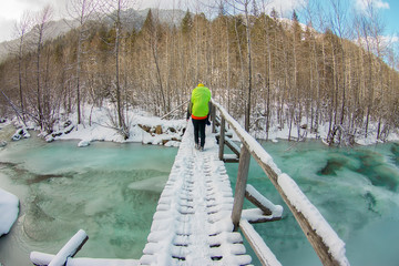 Girl backpacker walking on a bridge over a frozen turquoise ice river in the winter forest in the mountains