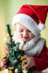 A toddler dressed as Santa Claus with a red cap on his head holds a small Christmas tree decorated with gold toys and looks at it.