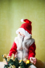 The kid in Santa Claus costume sits in a hat, stretched on the face next to a small Christmas tree.