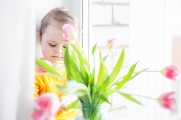 Obraz na płótnie Canvas A child in a yellow T-shirt sits on a window sill near a window behind a bouquet of tulips