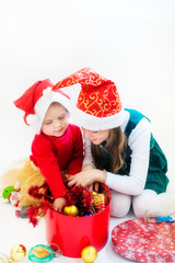 Two little girls in Santa Claus hats sit on their laps and take out Christmas toys from a big red box.