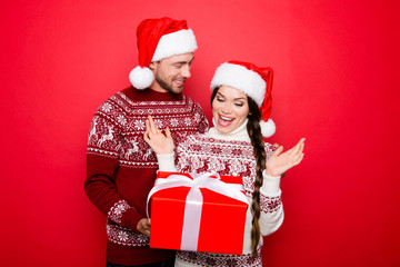 X mas promotion and miracle sale time! Cheerful partner in knitted clothing, head wear holds wrapped box, his astonished lady with raised palms of arms, she has braided brunette hair