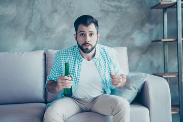 Excited man with bristle cheer for his favorite team, he is football fan, sitting on couch in casual outfit, holding a beer in hand