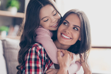 Playful small girl with long dark hair is hugging her mum's neck and looking at her with a smile, they have perfect weekends at home