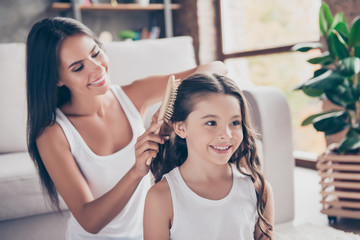 Close up photo of happy smiling schoolgirl with curly long dark hair and her beautiful mum who is brushing her hair, they are dressed in white tshirts