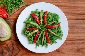 Fresh avocado, arugula and pepper salad. Green arugula and avocado salad with fresh pepper and sesame on a white plate and brown wooden table. Raw vegan meal. Top view 