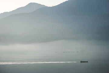 a small boat is sailing in the foggy lake in the morning with mountain backdrop. In the early morning at Sun Moon lake, a fisher man is sailing a boat to to find fish in the lake. The vintage photo. 