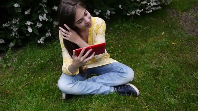 Beautiful teenager girl with tablet computer sits on the grass in Park. Movie of static camera.