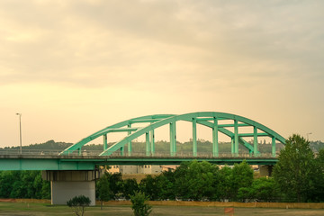 Green truss bridge under the sunset in the evening. The sky is so vanilla and calm. The steel metal bridge structure is strong. Vehicles can run on the bridge to cross a river underneath.   