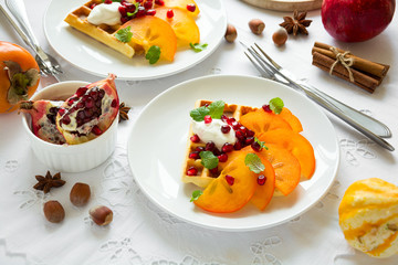 Plates of Belfian waffles with persimmon, pomegranate seeds and sour cream on white table cloth