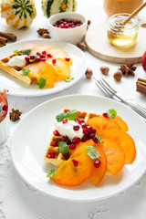 Plates of Belgian waffles with persimmon, pomegranate seeds and sour cream on white table cloth