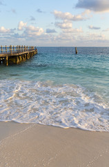 Morning at the Caribbean shore. Long wooden pier on the left. Foamy waves and turquoise water. Peaceful scenery. Wallpaper.
