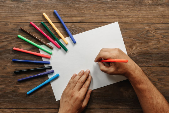 Hands of the artist close-up, holds a flamaster over a blank sheet of paper near scattered colored felt-tippers on a brown wooden table. View from above. Copy space.