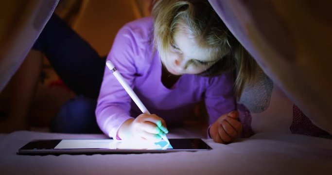 Child blonde caucasian girl digital coloring with tablet app under bed blanket at night. Modern technology childhood kid use at home. 4k video