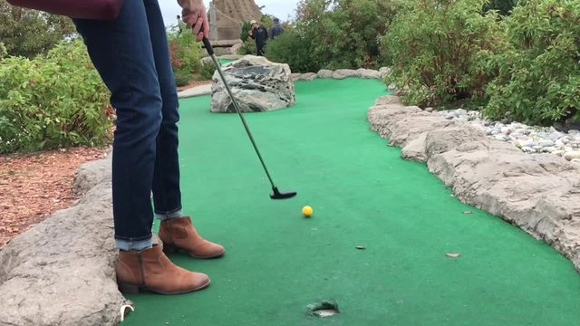 Lower part of woman wearing boots hits yellow golf ball in slow motion on mini golf course.  