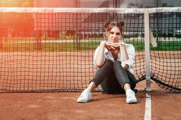 A beautiful girl in a white shirt, black pants and white sneakers sits at the net on the tennis court.
