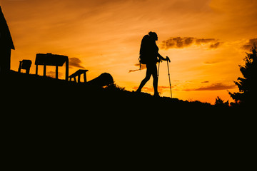 Silhouette hiker woman walking with backpack and trekking pole, sunset orange sky on the background
