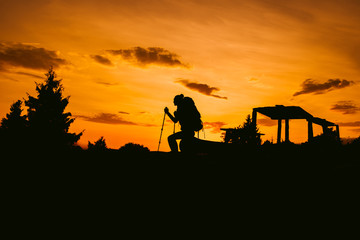 Silhouette hiker woman sit on the bench, with backpack and trekking pole, sunset orange sky on the background