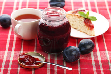 Plum jam in jar with cup of tea and cake on red napkin